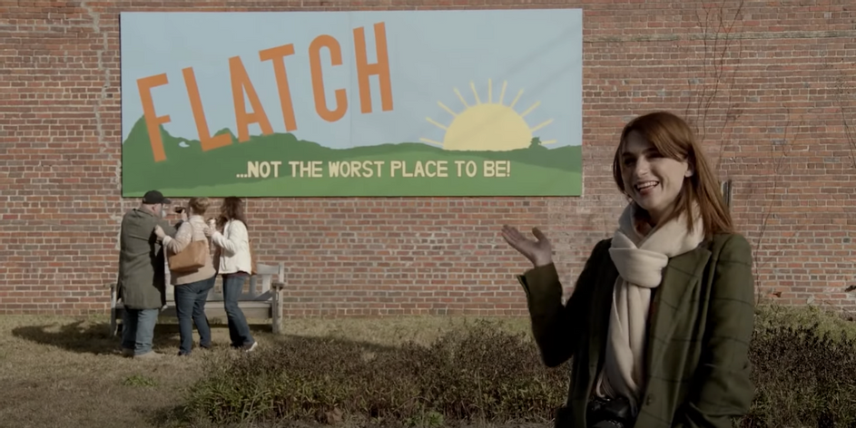 red haired female character gestures toward brick wall with sign that reads flatch not the worst place to be 
